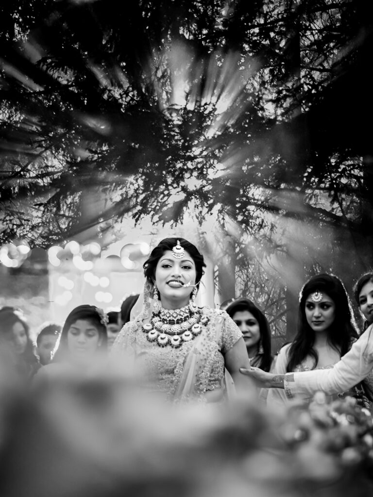 Black and white photo of a bride walking through a sunlit garden aisle