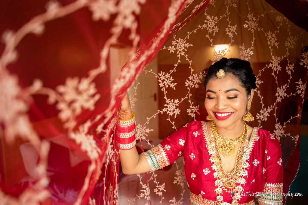 Bride smiles while reaching for a red draped curtain wearing gold jewelry
