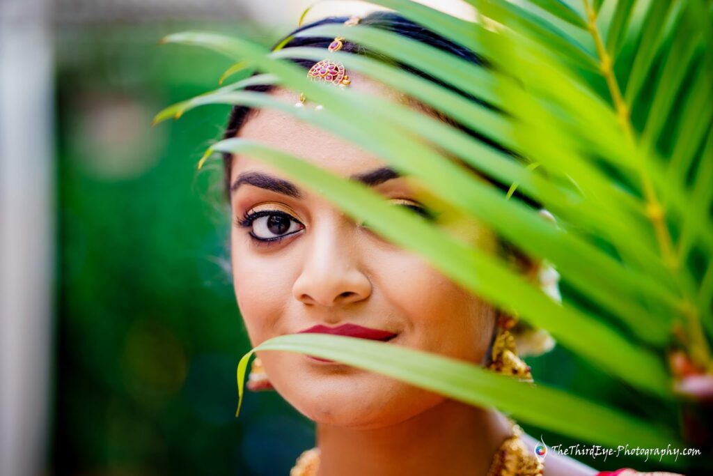A bride peeks from behind a green palm leaf outdoors