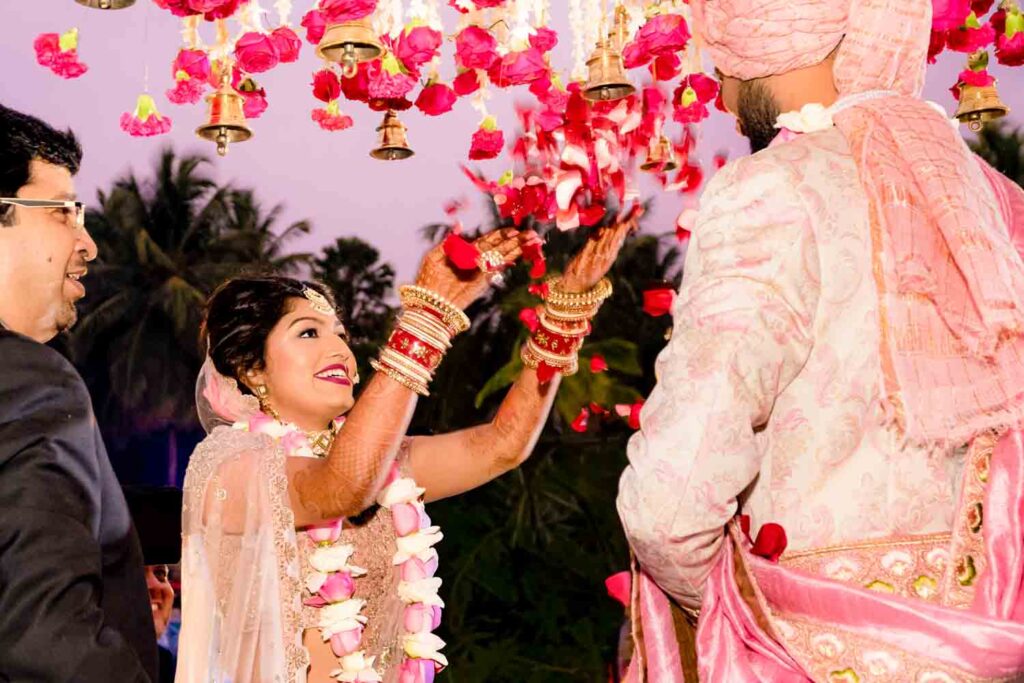 Bride lifts a floral garland toward the groom under a hanging flower canopy at dusk