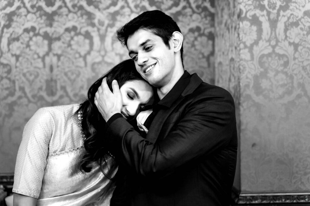 Black and white photo of a couple embracing against patterned wallpaper