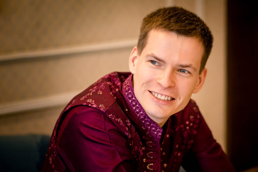 Groom smiles indoors wearing a maroon embroidered sherwani