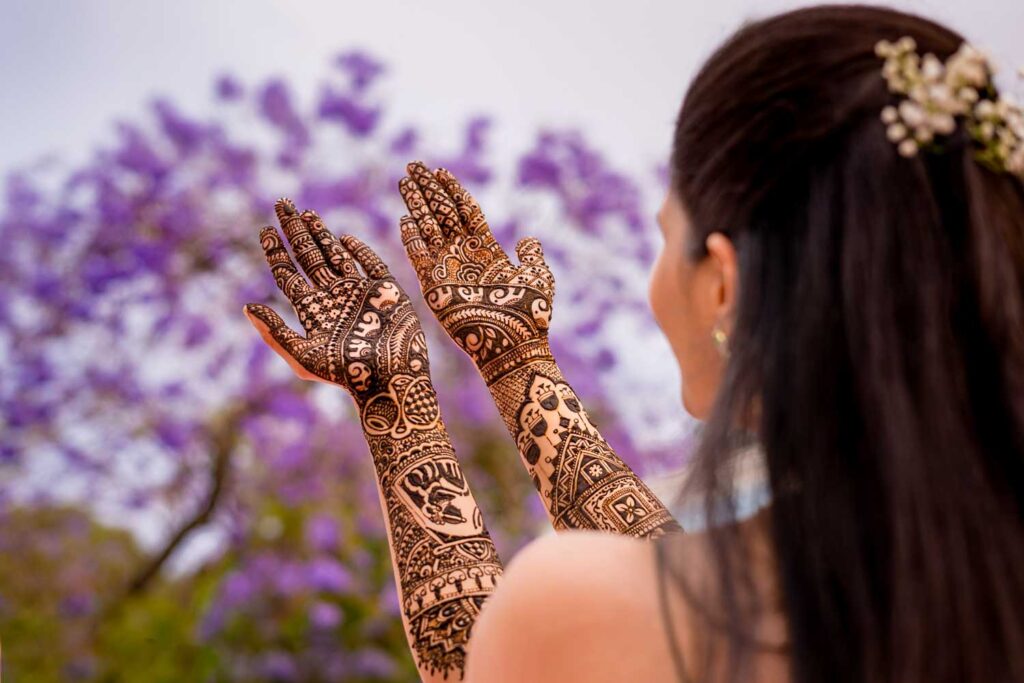 Bride raises mehndi covered hands against blooming purple jacaranda trees