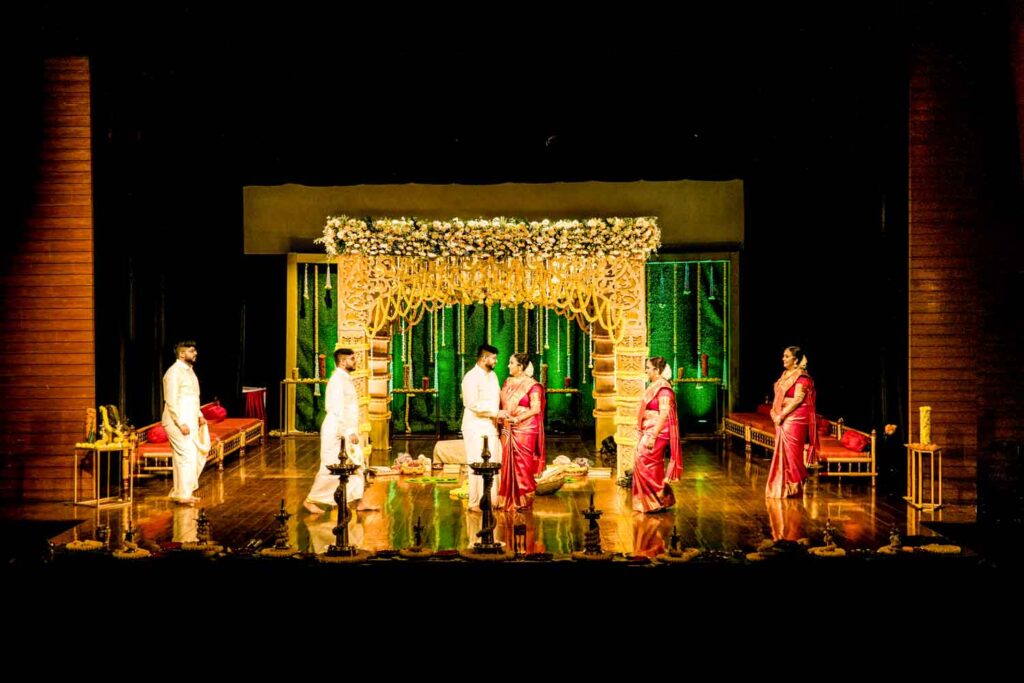 Priests and a couple perform wedding rituals on stage under a floral mandap