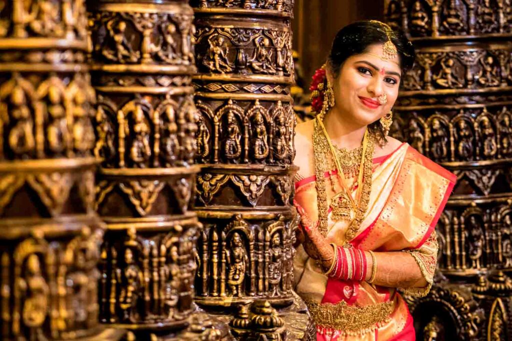 Bride in a red and gold saree stands with folded hands at an ornate carved temple