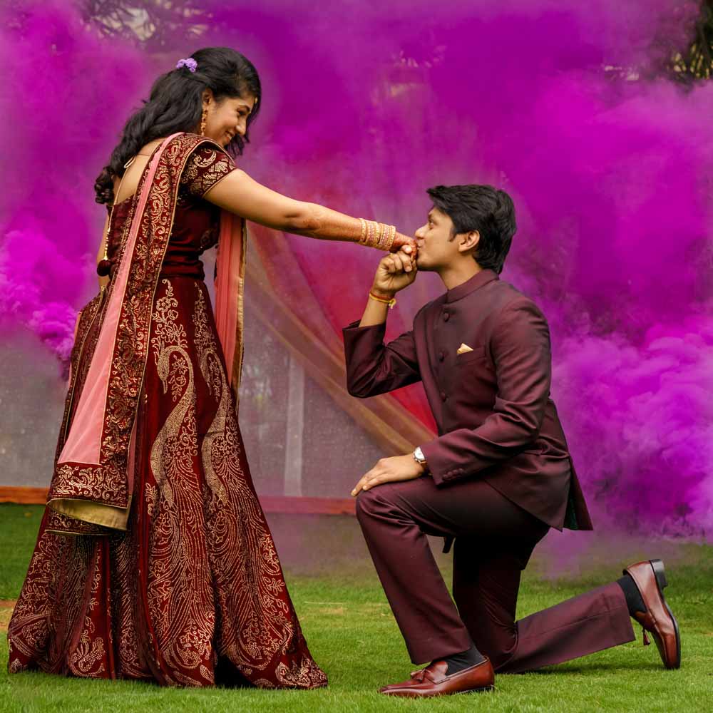 Groom kneels on the grass holding the bride close amid pink smoke