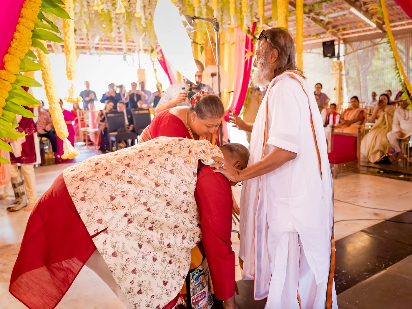 A person in a red and white kurta bends low to touch the feet of a man in white robes