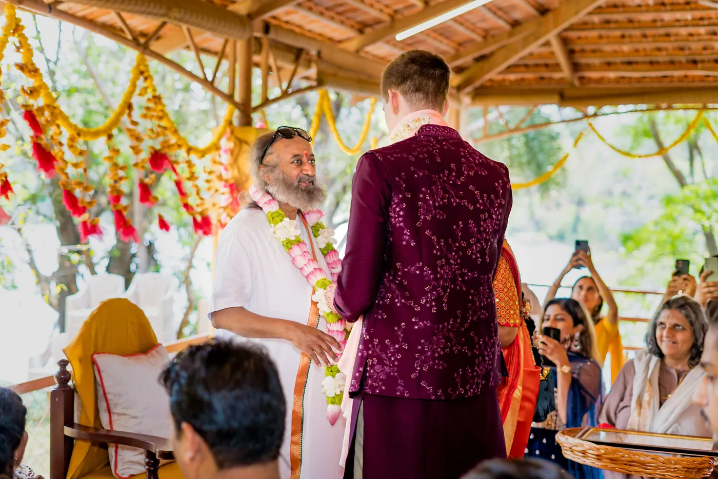 Stefan in a magenta jacket faces Sri Sri Ravi Shankar in white robes as a woman holds a tray
