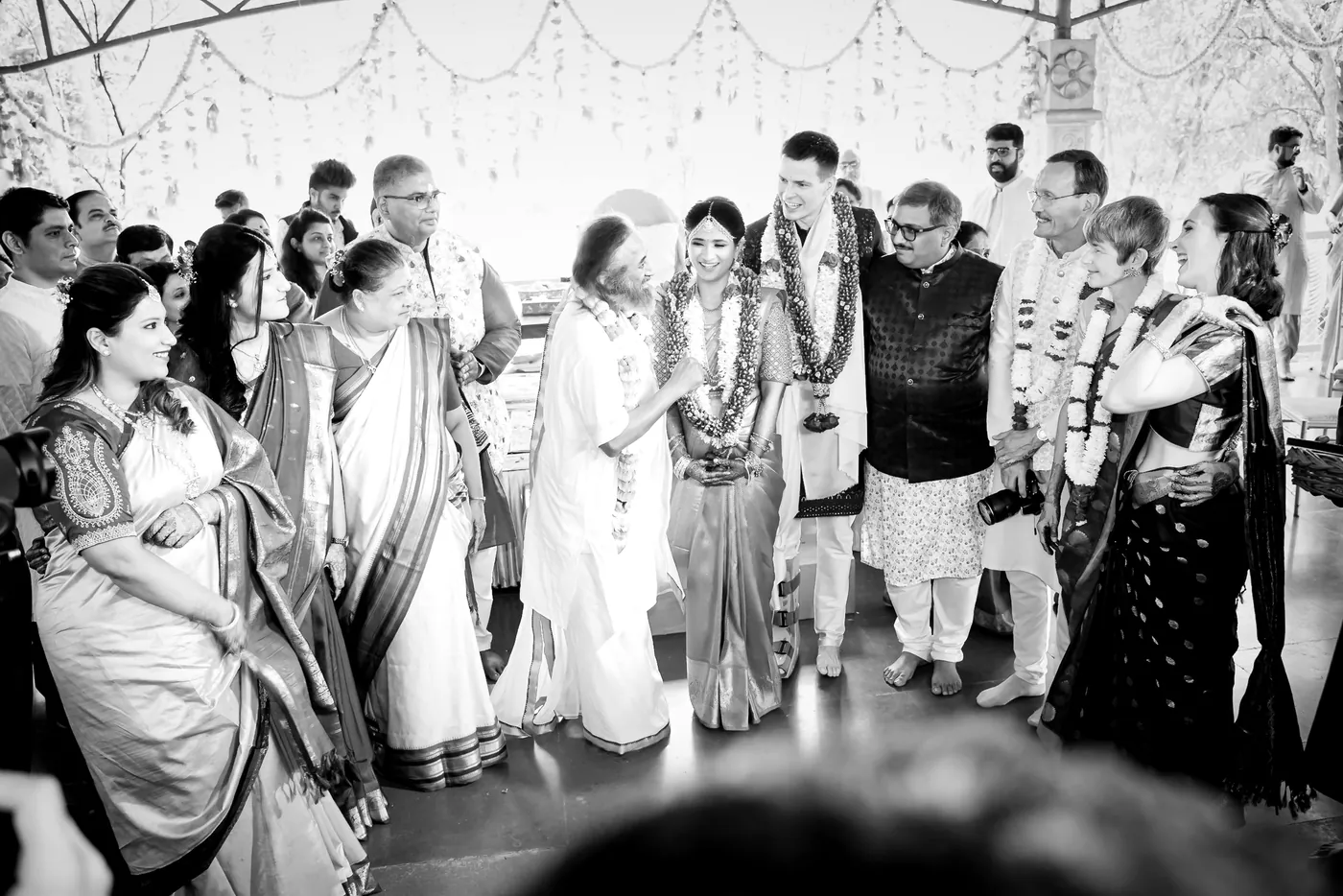 Black and white of the couple standing among guests and family around Sri Sri Ravi Shankar