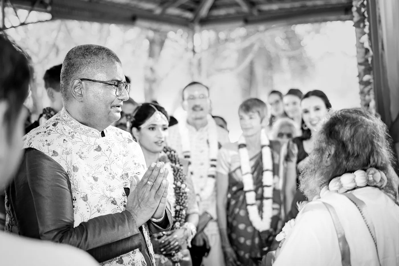 Black and white of a man with folded hands looking toward Sri Sri Ravi Shankar and the couple