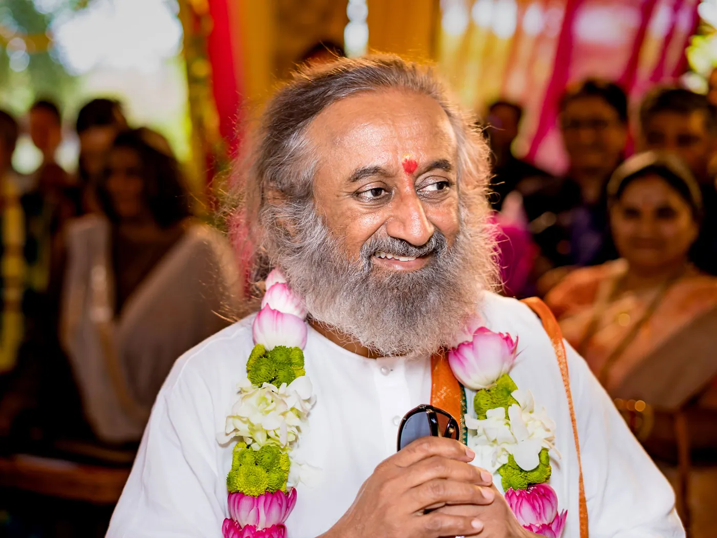 Sri Sri Ravi Shankar smiles holding a microphone and a garland of pink lotus flowers