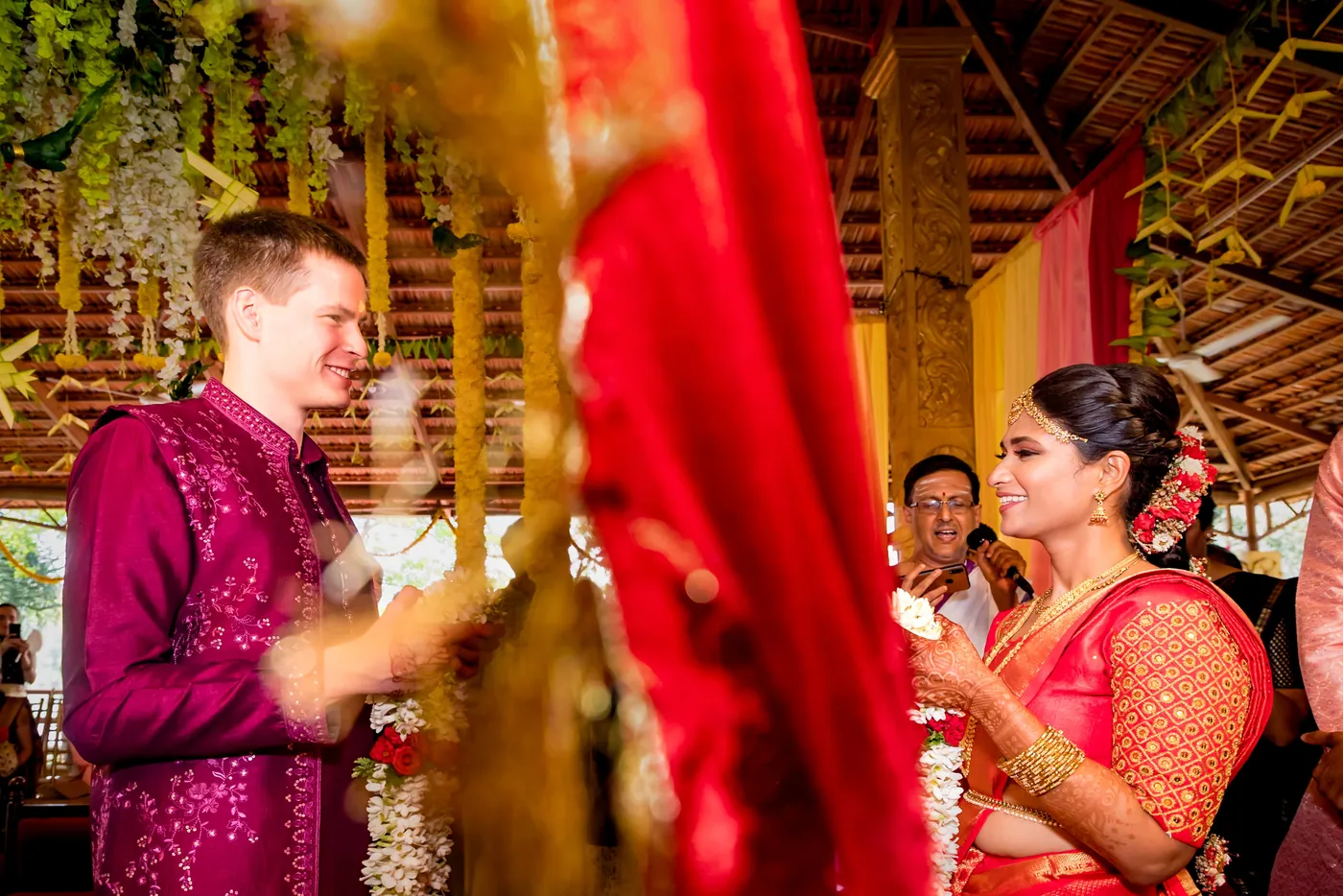 Stefan in a magenta embroidered jacket stands near Medha in an orange blouse beside a red drape at the mandap