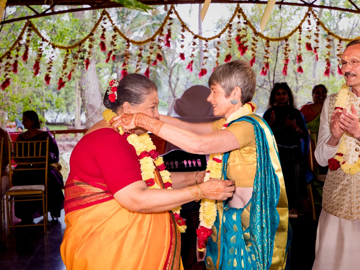 A woman in an orange sari garlands a guest in a teal sari under hanging flower strings