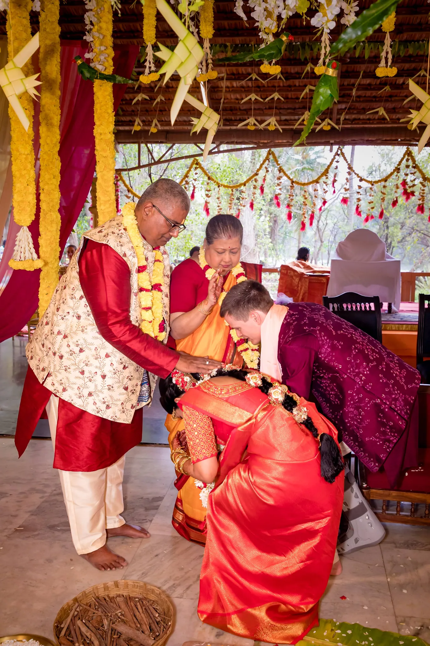 Medha and Stefan bend low to touch the feet of an elder in a cream waistcoat under marigold garlands