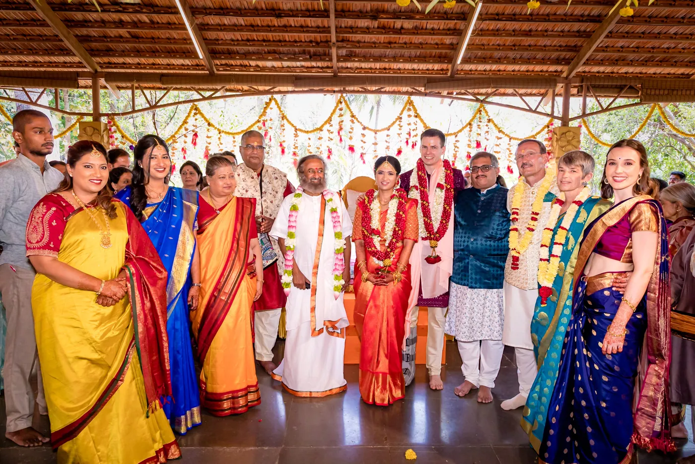 Medha and Stefan stand with a large group of family and a priest in white at the ceremony hall