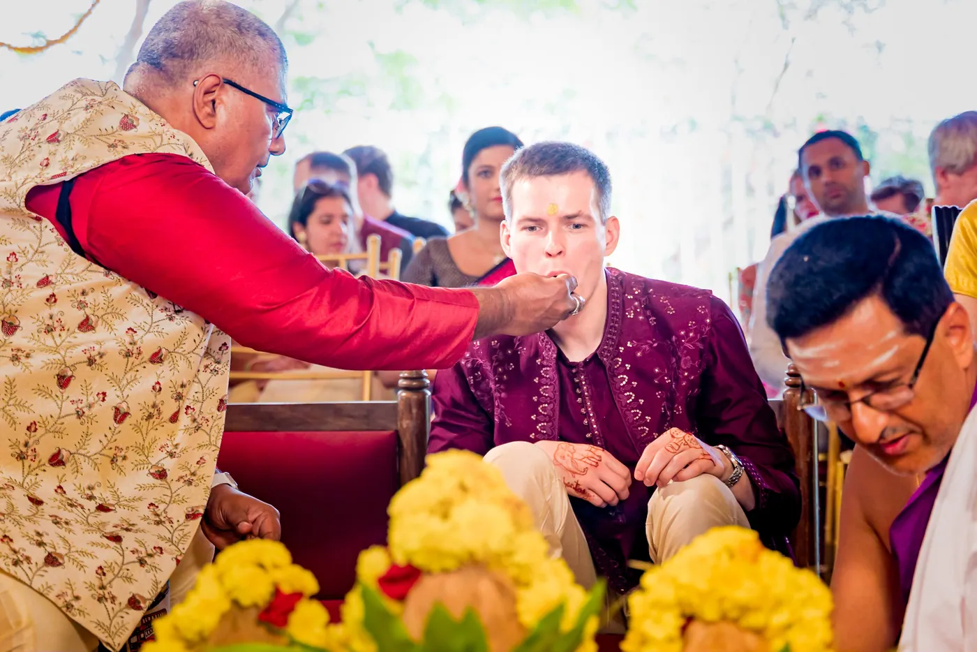 A man in a red and gold jacket offers something to Stefan while a priest sits beside them