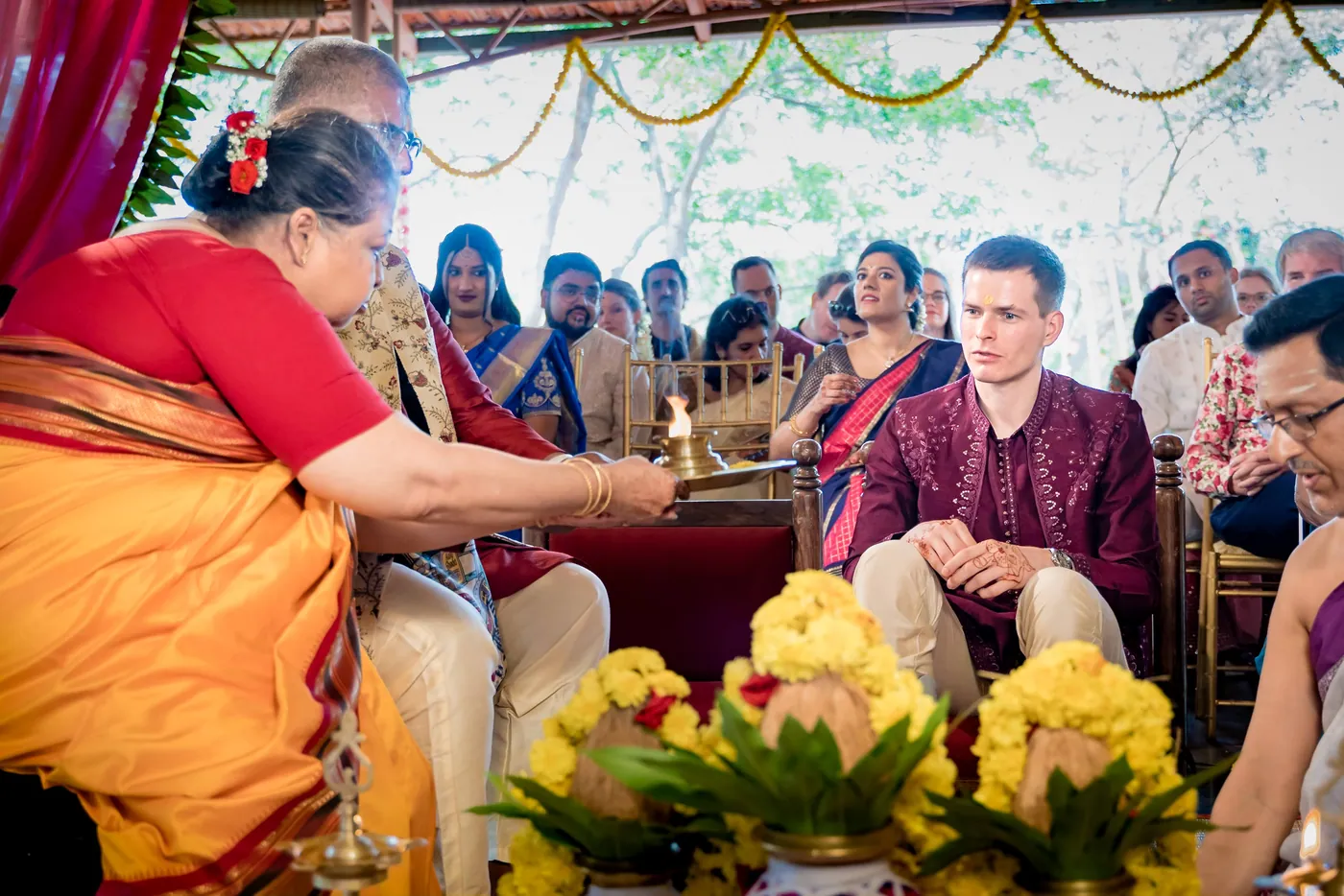 A woman in a red sari lights a lamp while Stefan sits behind the flowers