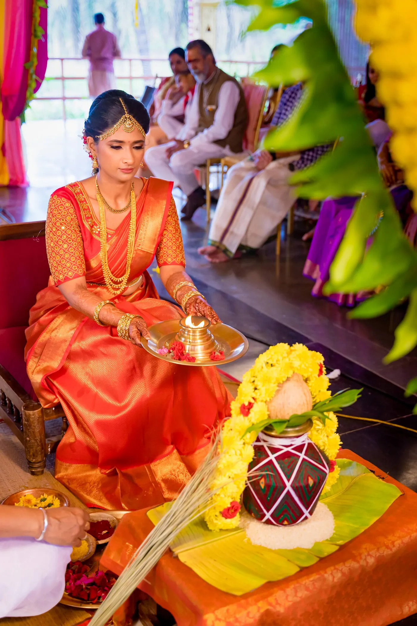 Medha in an orange sari sits cross-legged performing a ritual with a plate and decorated pot