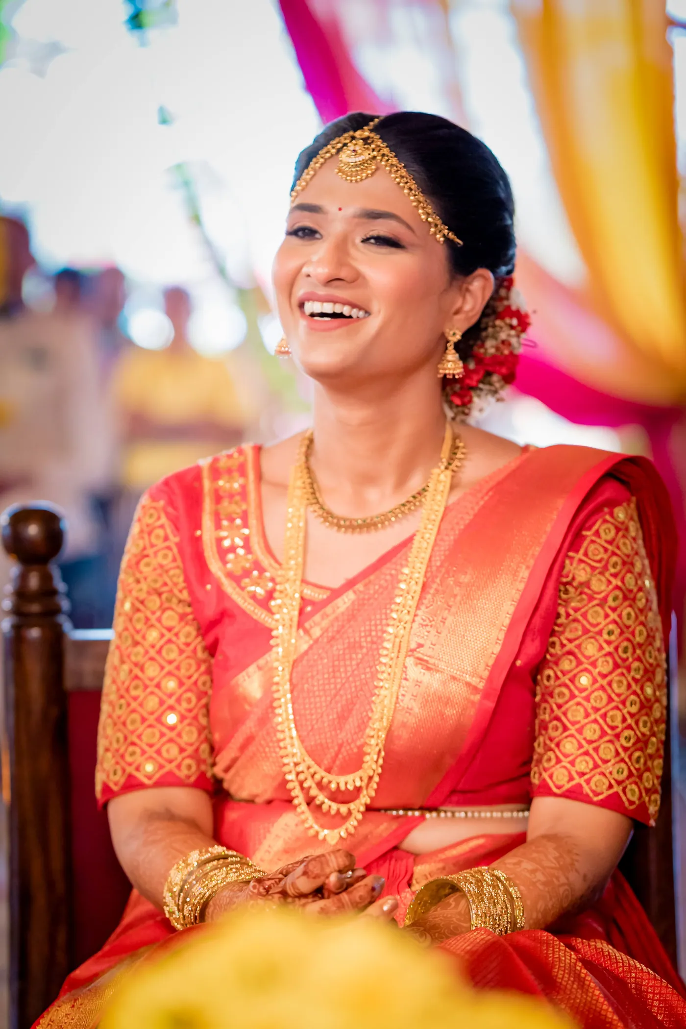 Medha laughs joyfully in an orange sari and gold necklace on a carved wooden chair