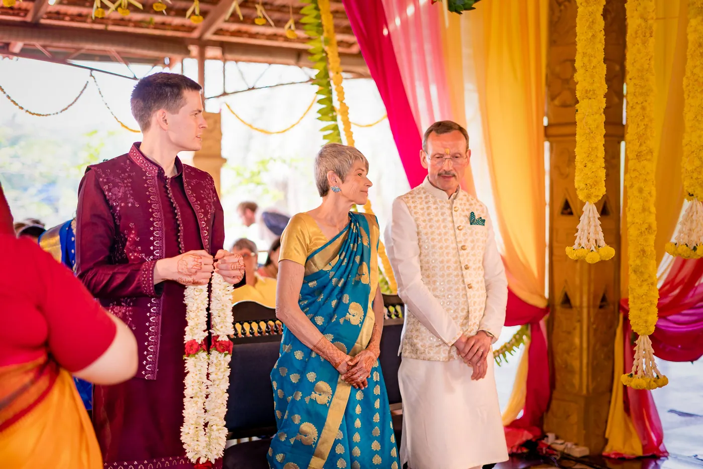 Stefan holds a flower garland beside a woman in teal and a man in cream before the ceremony
