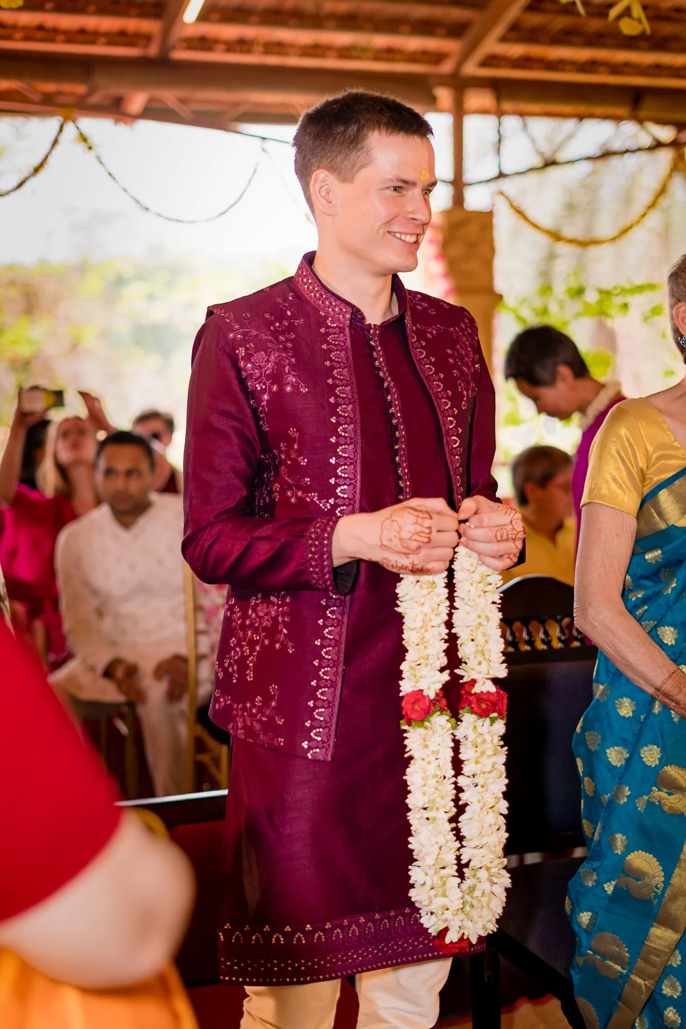 Stefan stands in a magenta embroidered jacket holding a garland of white jasmine flowers