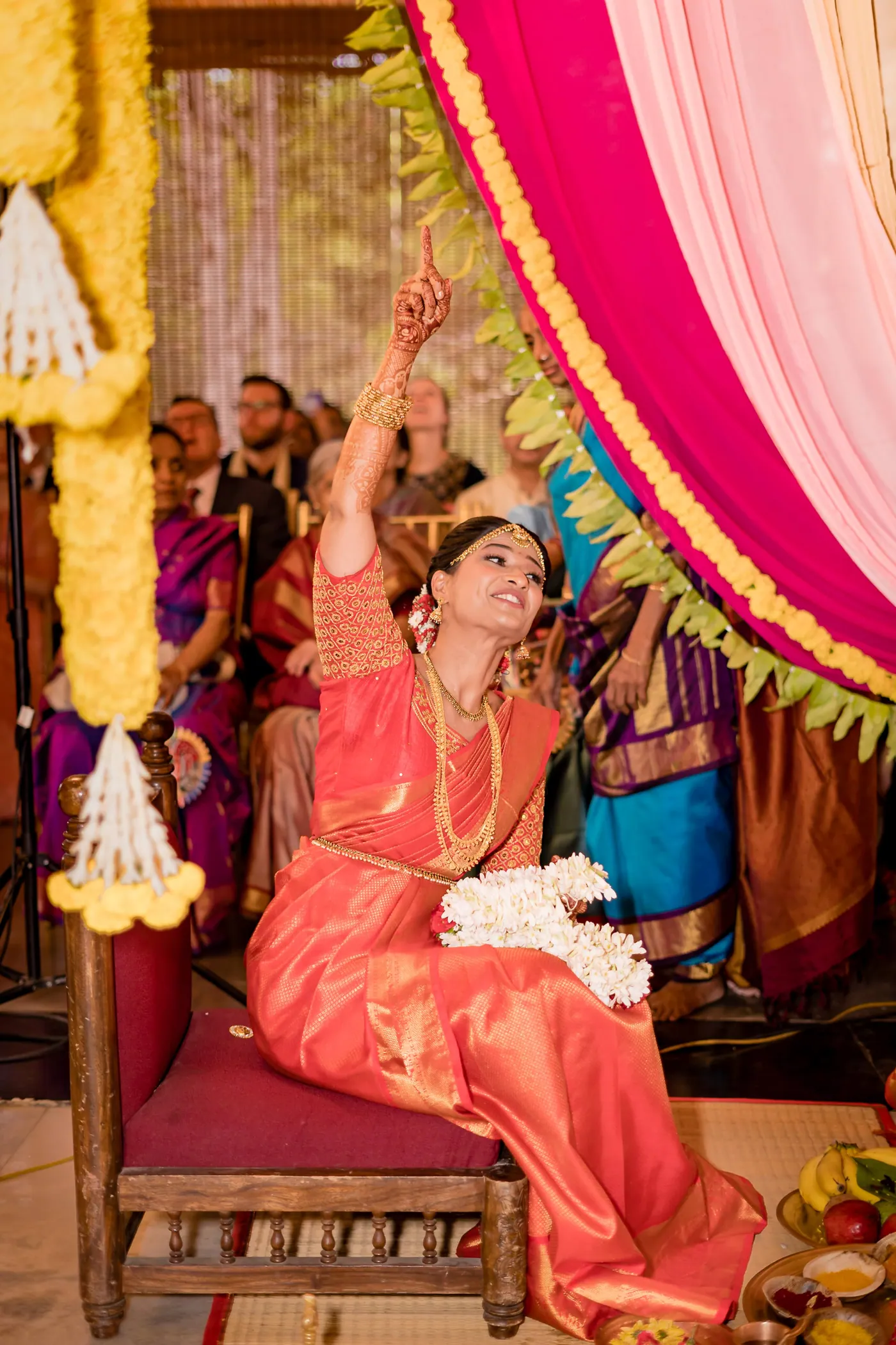 Medha sits on a carved chair in orange raising her arm in celebration