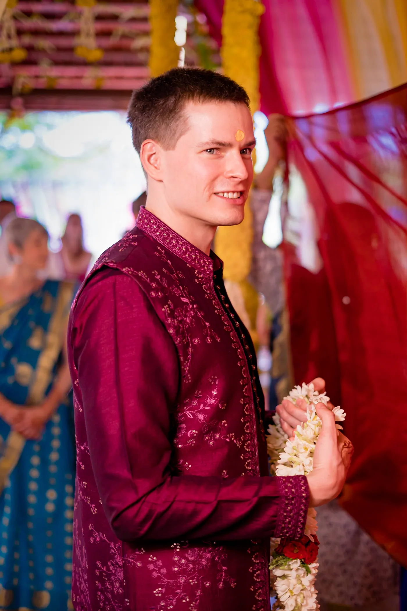 Stefan in a magenta jacket smiles while holding white flower garlands beside a red curtain