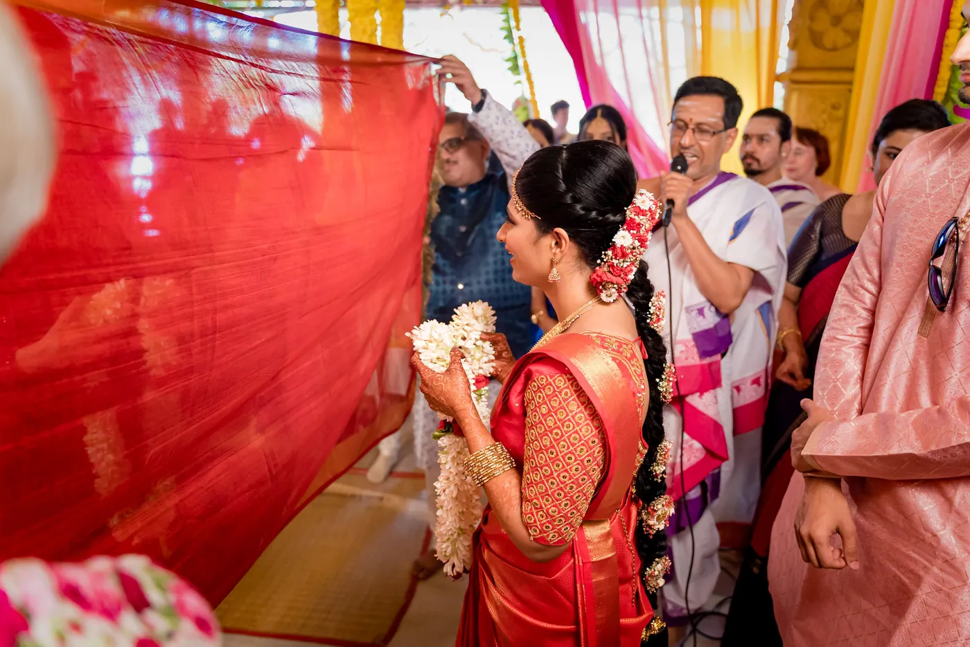 Medha holds white flowers behind a red cloth held up by guests crowding close