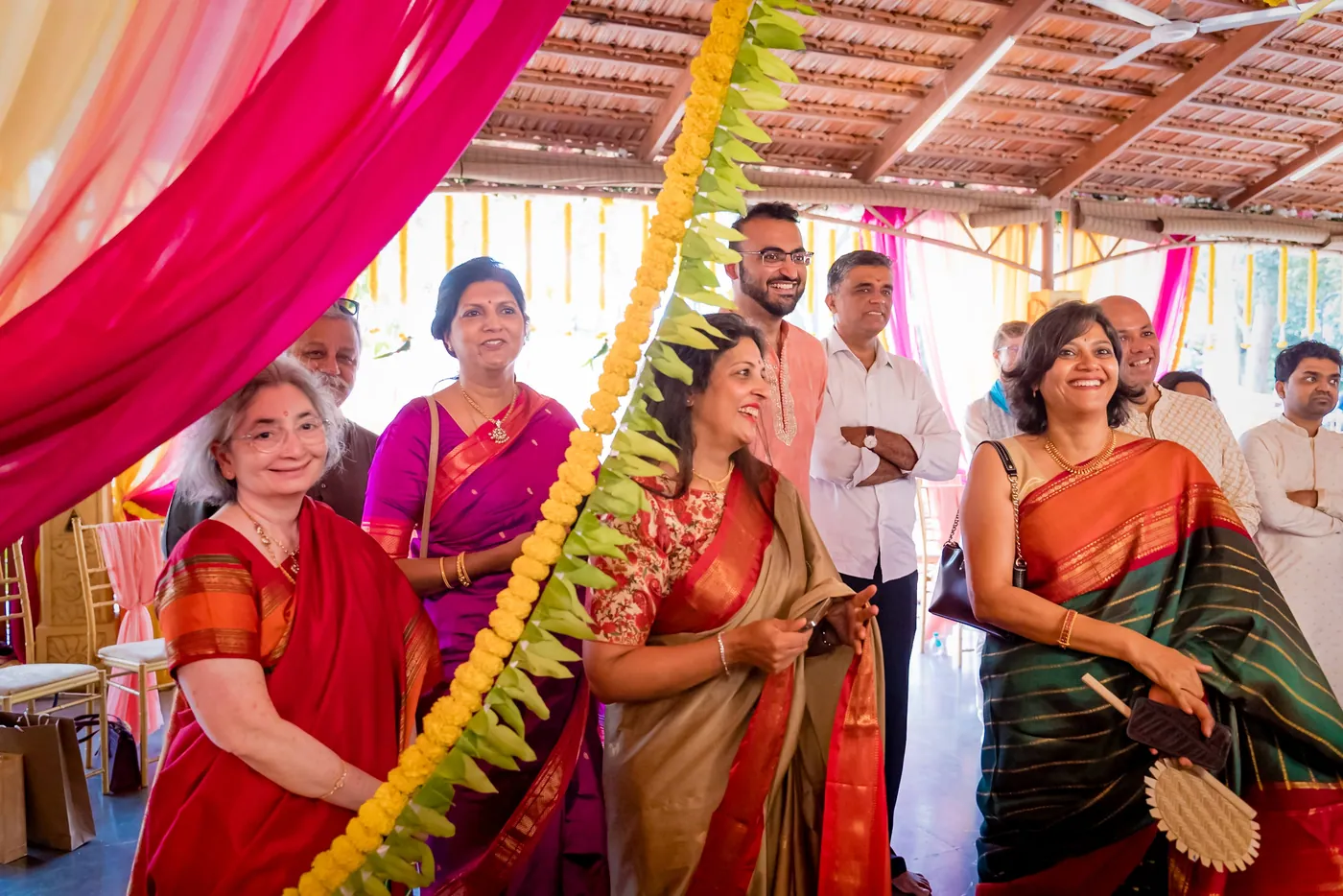 Women in bright saris smile and watch under hanging flowers during the ceremony