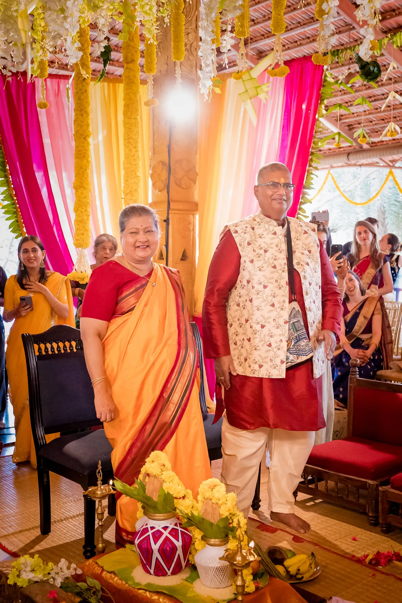 A woman in an orange sari and a man in a cream waistcoat stand before the audience