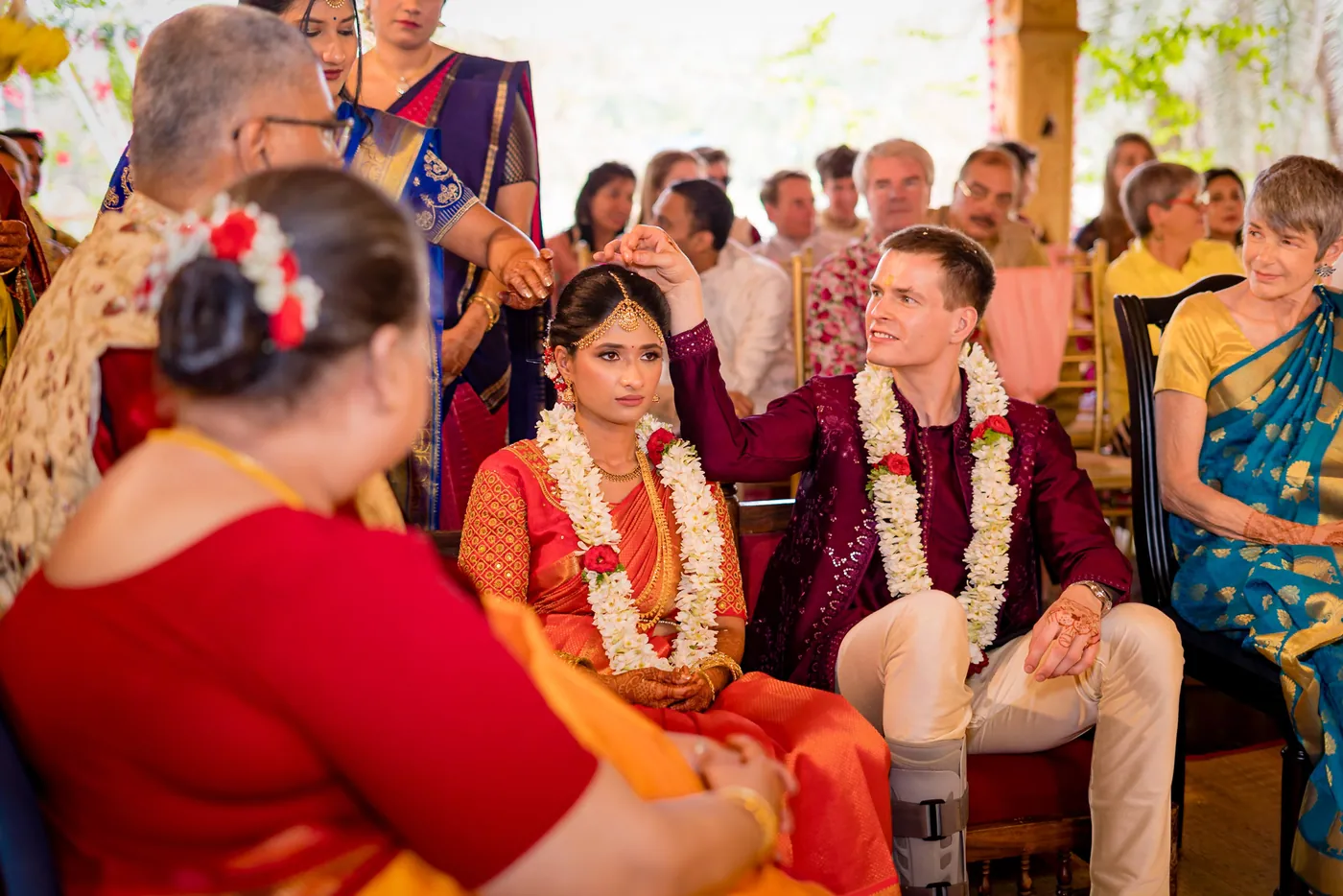 Medha and Stefan sit together in garlands as an older woman talks with them