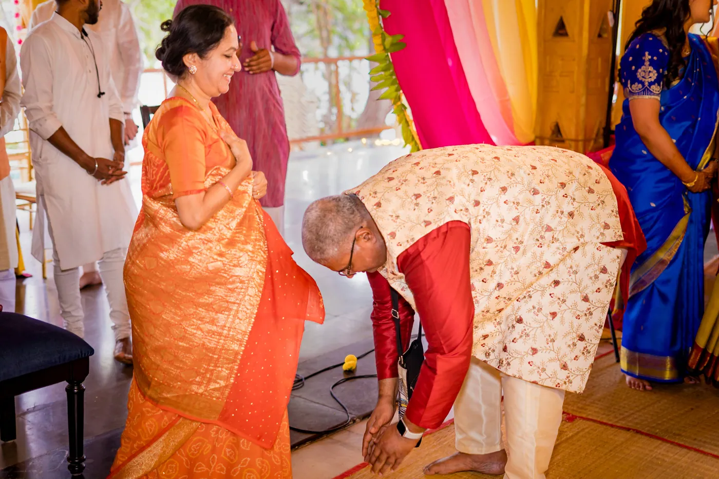 An older man in a cream waistcoat bends to touch the feet of a woman in an orange sari