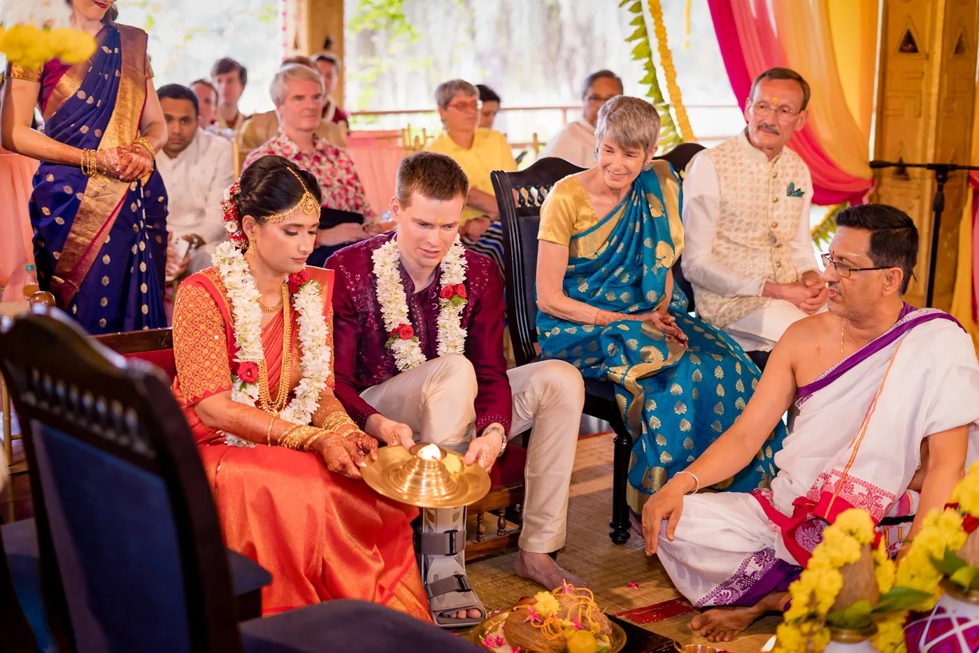 Medha and Stefan sit before a priest and guests, with a small brass dish before them