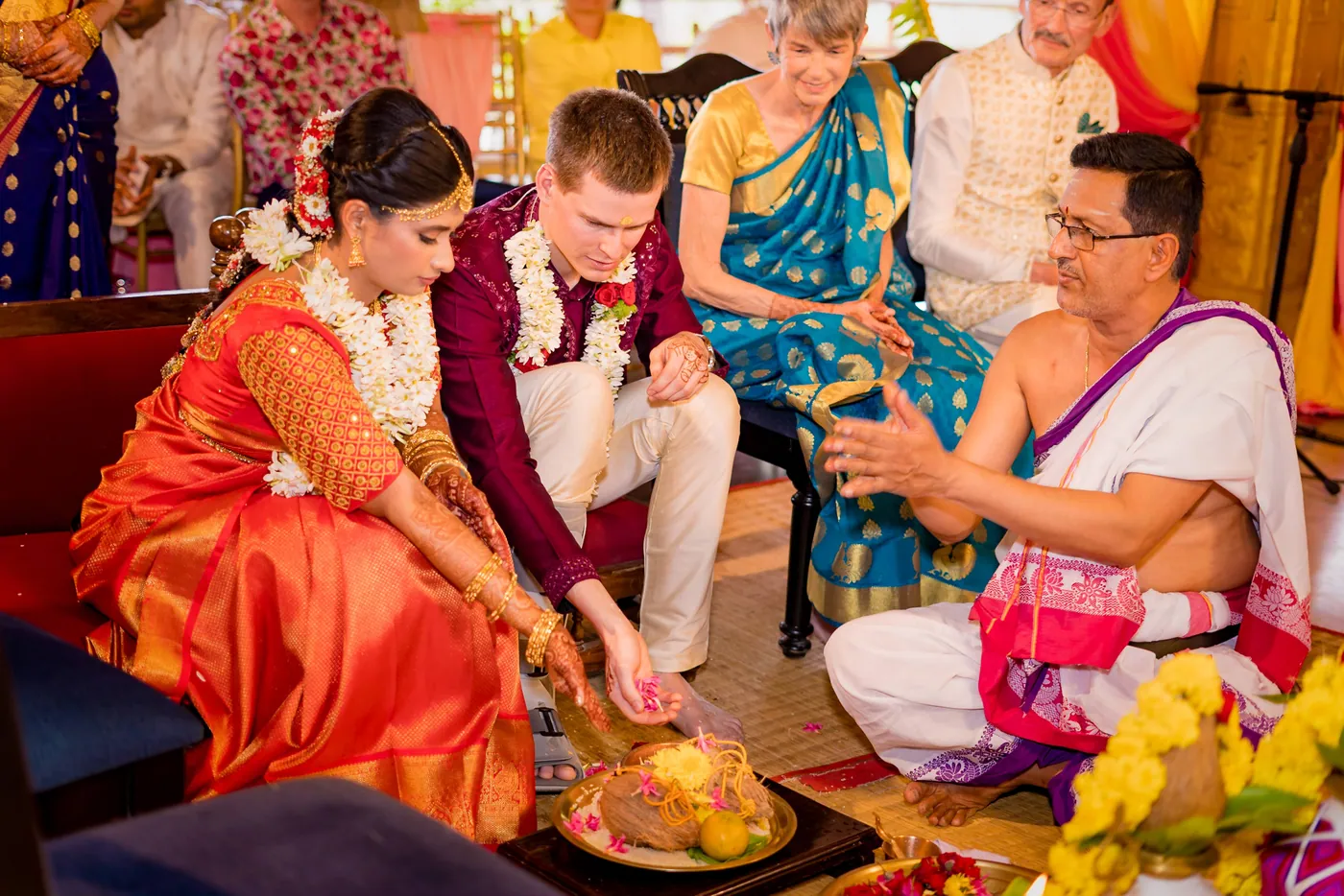 Medha and Stefan perform a ritual with a priest in white beside a brass dish