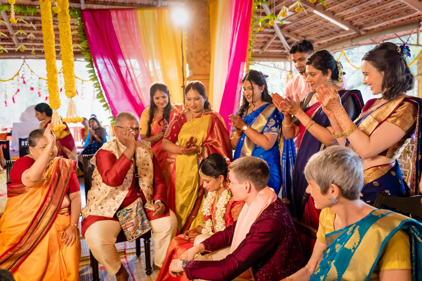 Family members in bright saris and waistcoats gather around Medha and Stefan, applying marks
