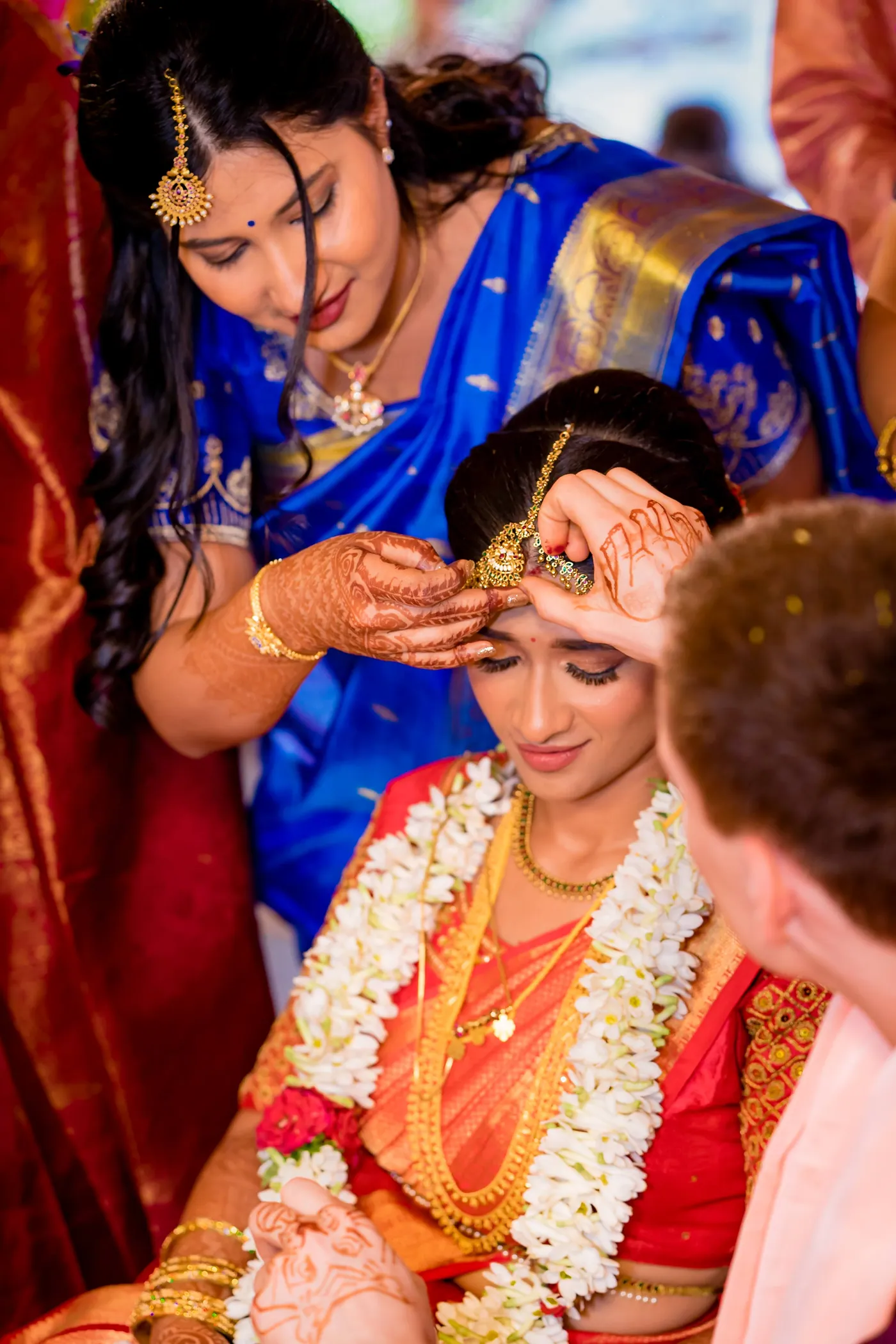 A woman in blue applies a mark on Medha's forehead while Stefan watches