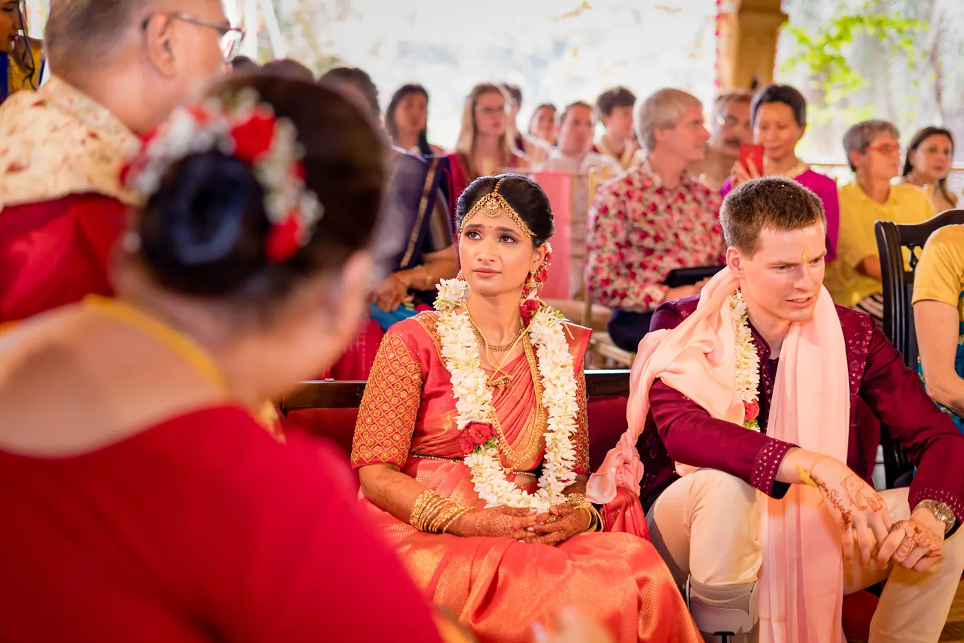 Medha and Stefan sit together smiling in garlands as family gathers around them