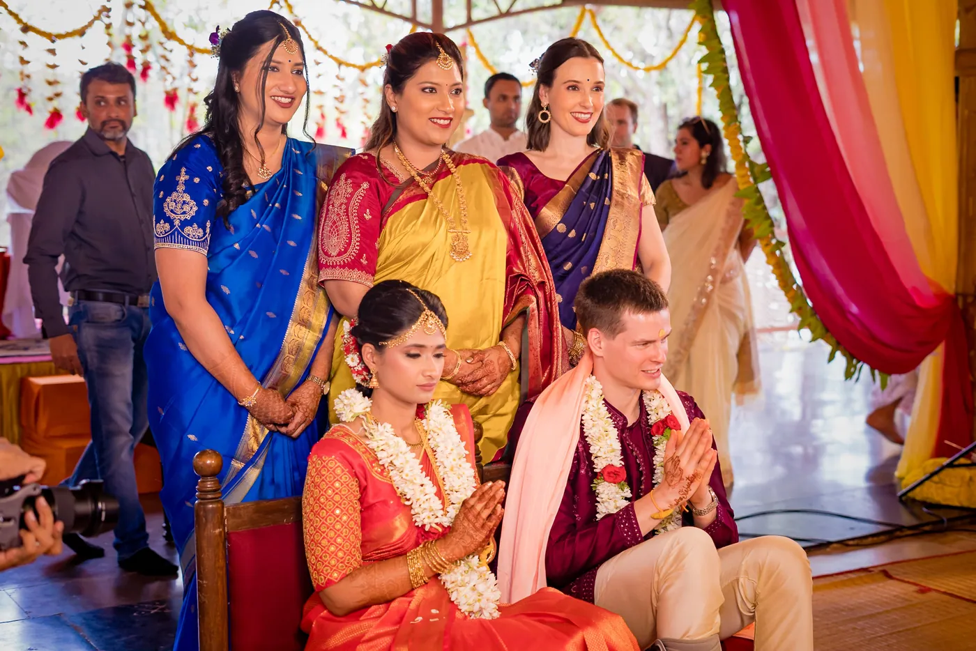 Medha and Stefan sit with folded hands as women in saris smile behind them