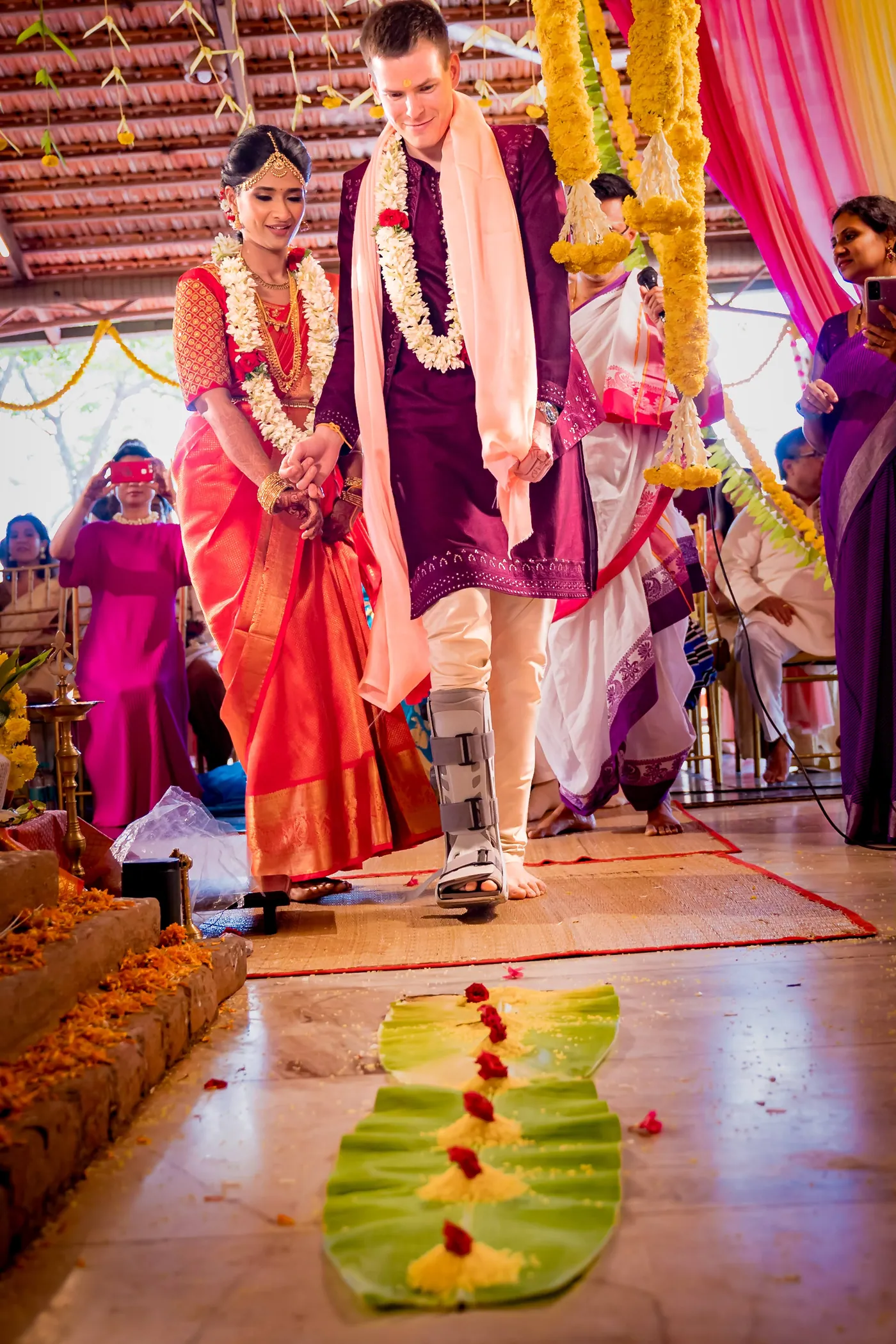 Medha steps over banana leaves with red flowers while Stefan walks beside her in a walking boot
