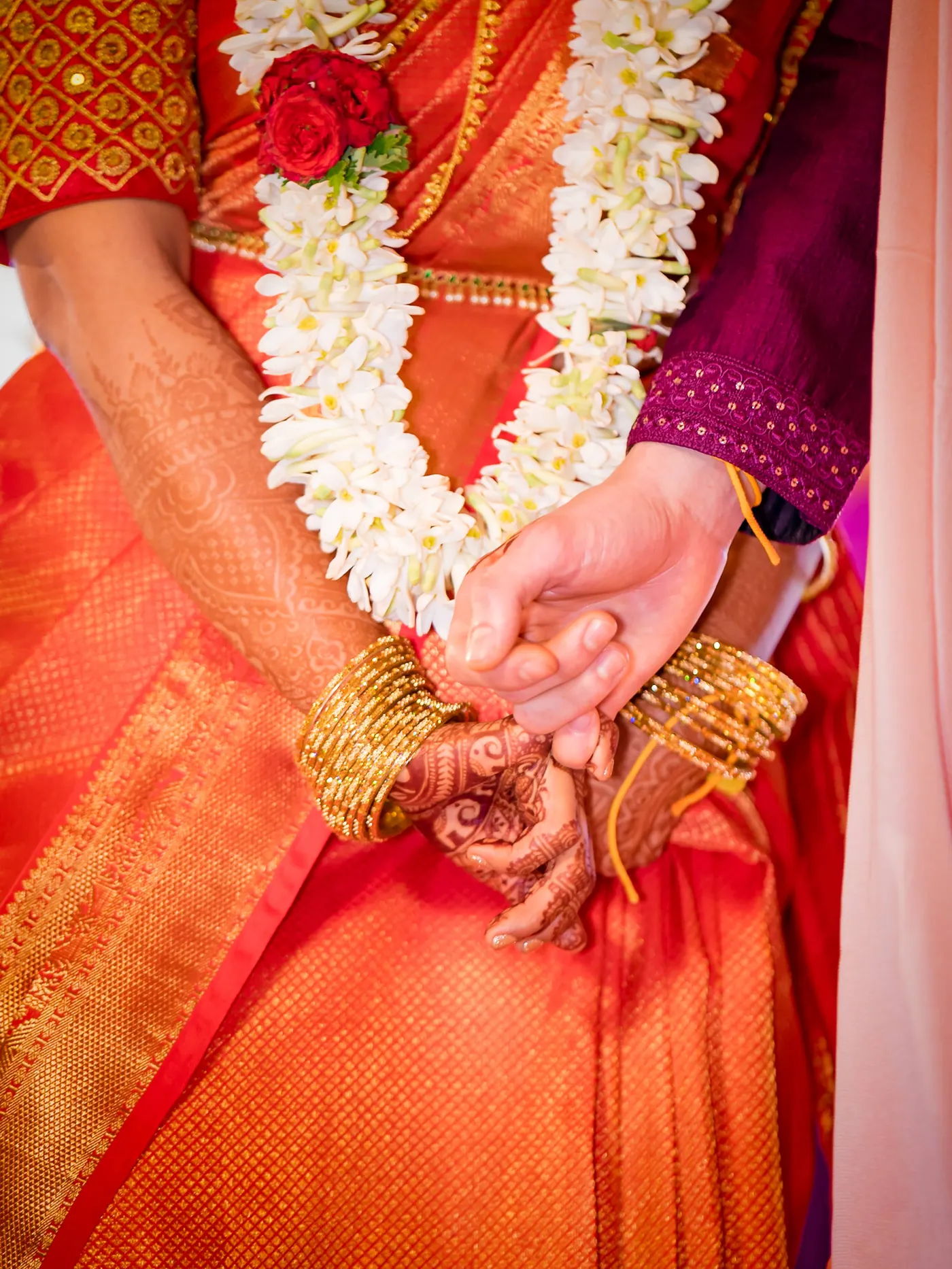 Medha and Stefan hold hands wearing white garlands over orange silk and a magenta jacket