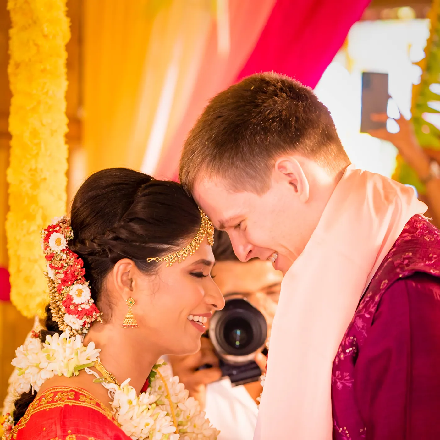 Medha and Stefan touch foreheads and smile as a photographer leans close