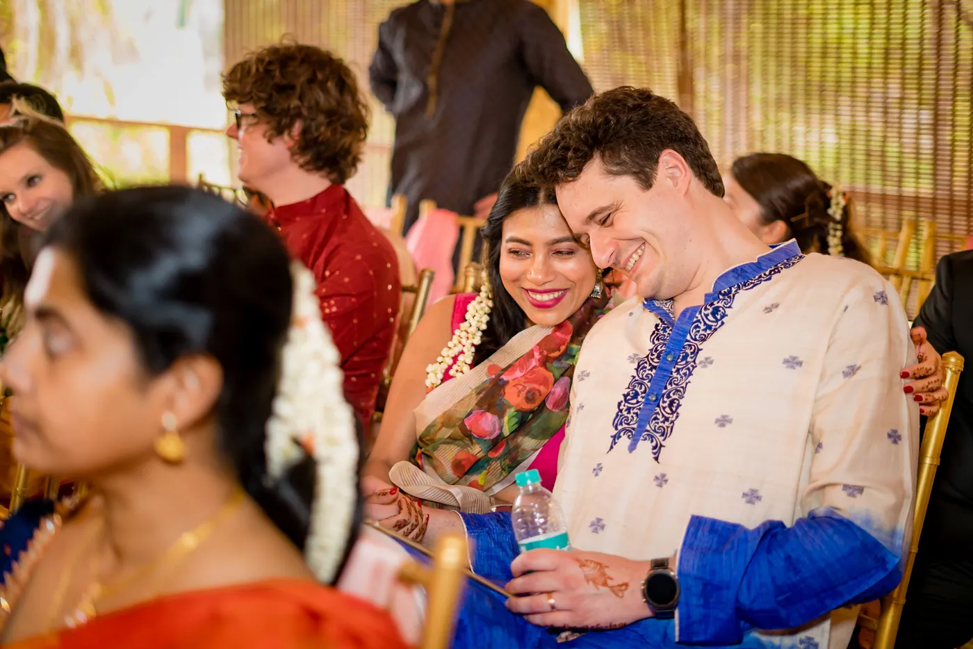 A smiling guest in a blue and white kurta sits close with a woman beside Medha and Stefan