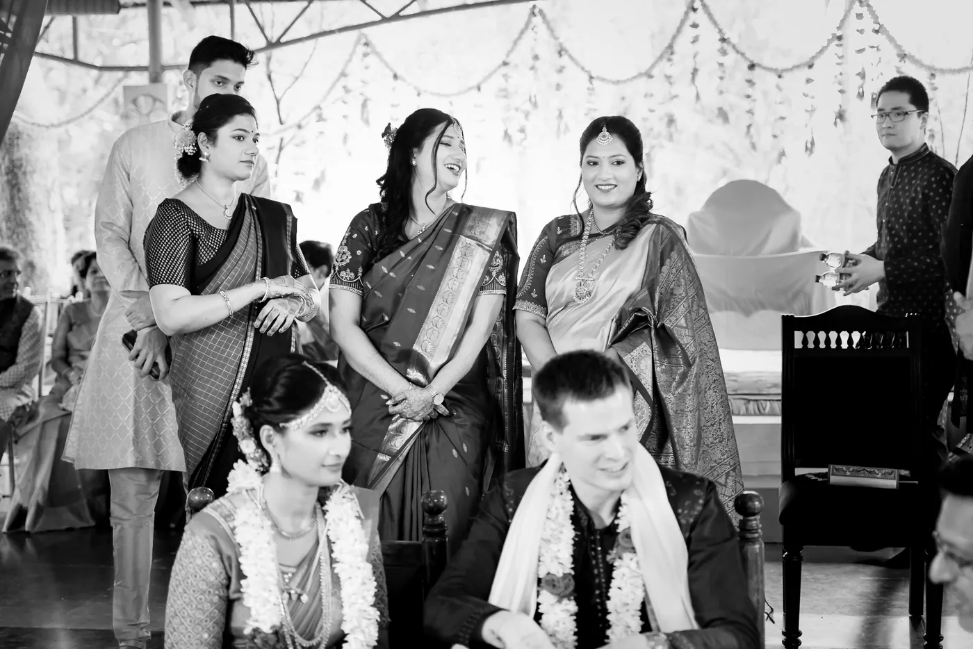Black and white of Medha and Stefan sitting with guests in saris behind them