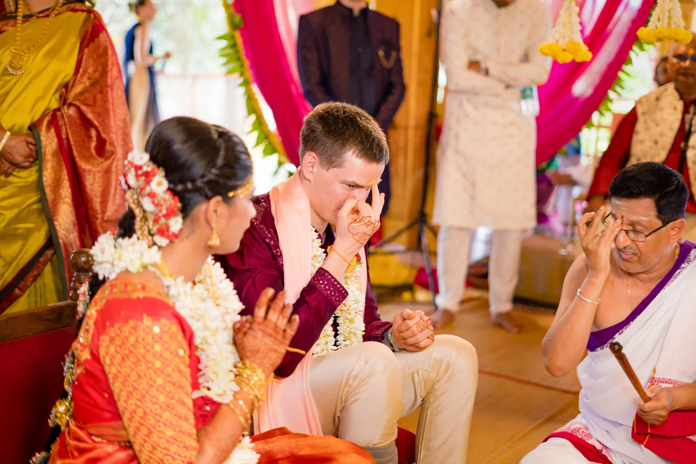 Medha smiles beside Stefan, who wipes his eye, as a priest and elders look on
