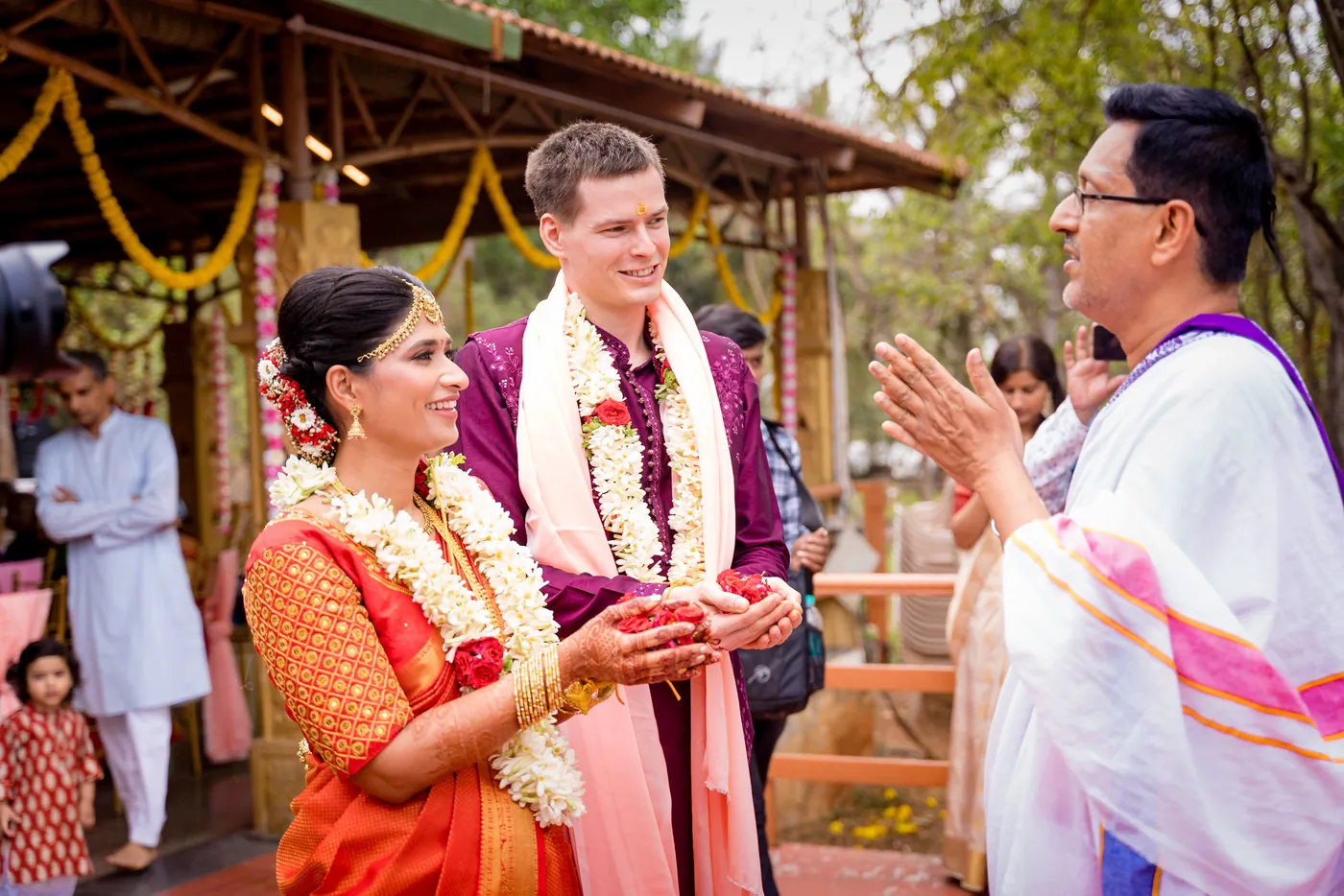 Medha and Stefan hold flower petals and smile as a priest in white speaks with them