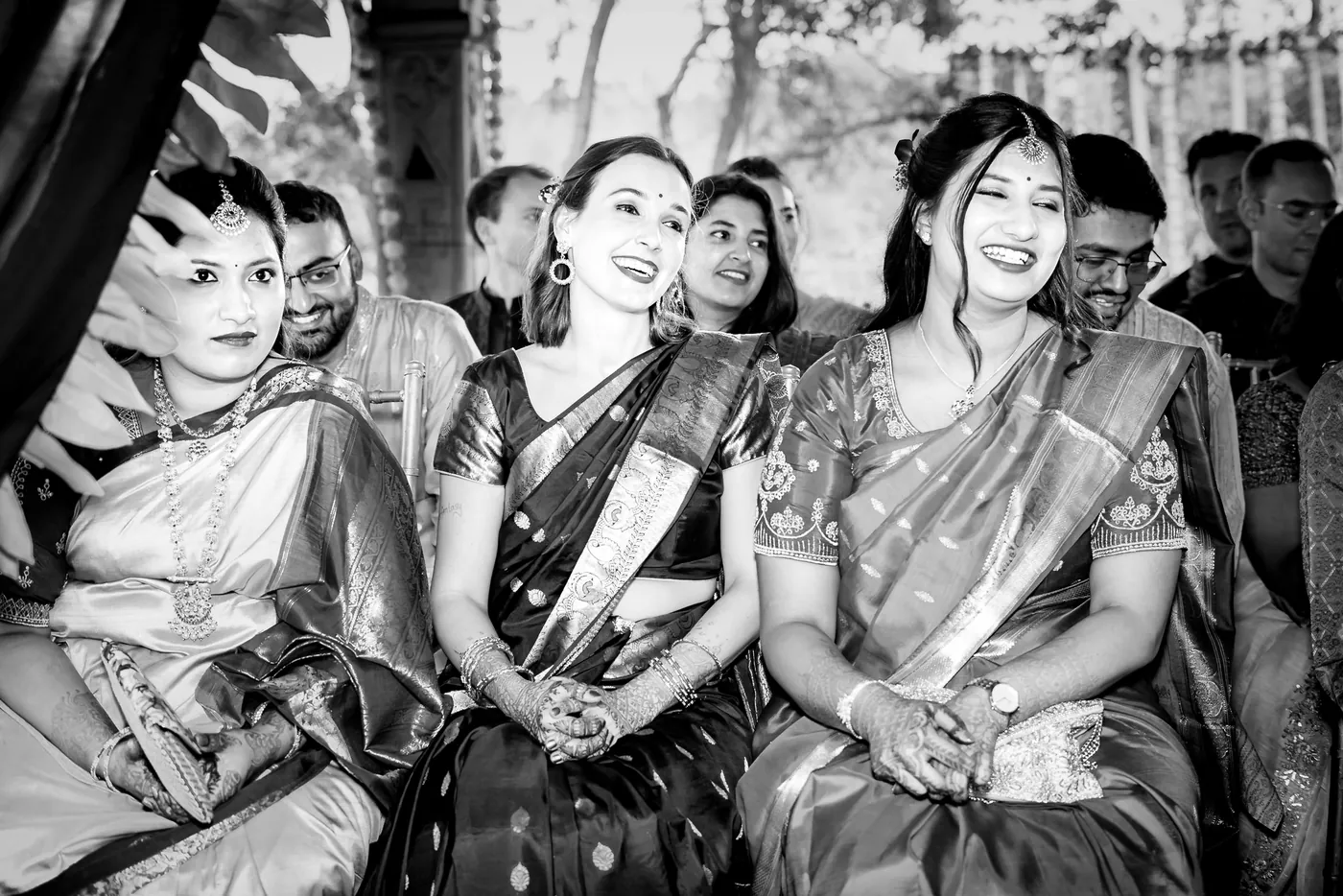 Black and white of Stefan's sister laughing among seated guests in saris with her hands clasped