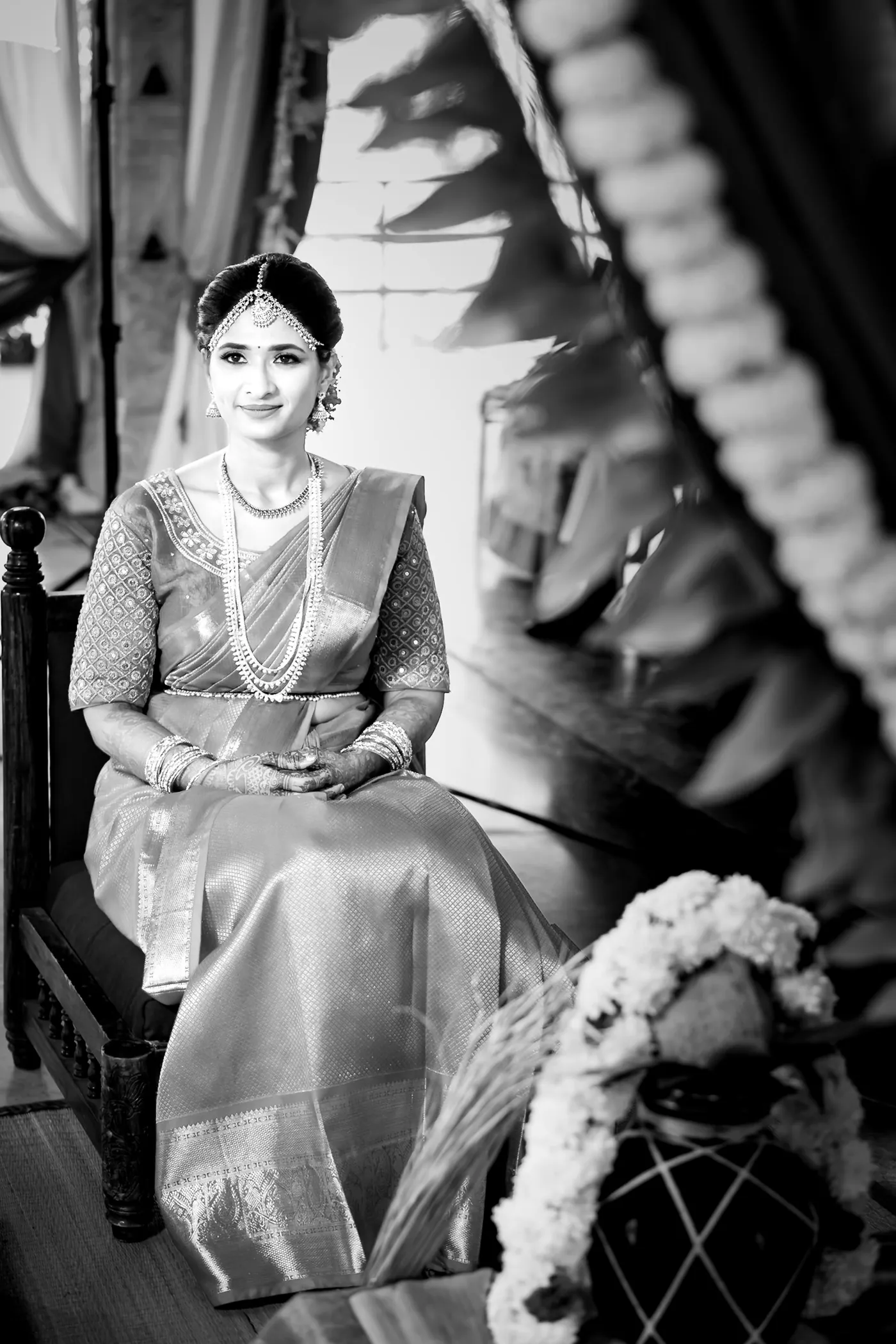 Black and white of Medha seated on a carved wooden chair with a white flower garland beside her