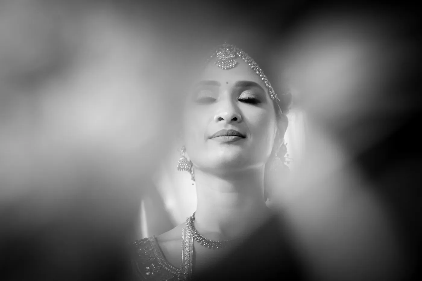Black and white of Medha, eyes lowered and a gold headpiece, seen through a soft blur of foreground