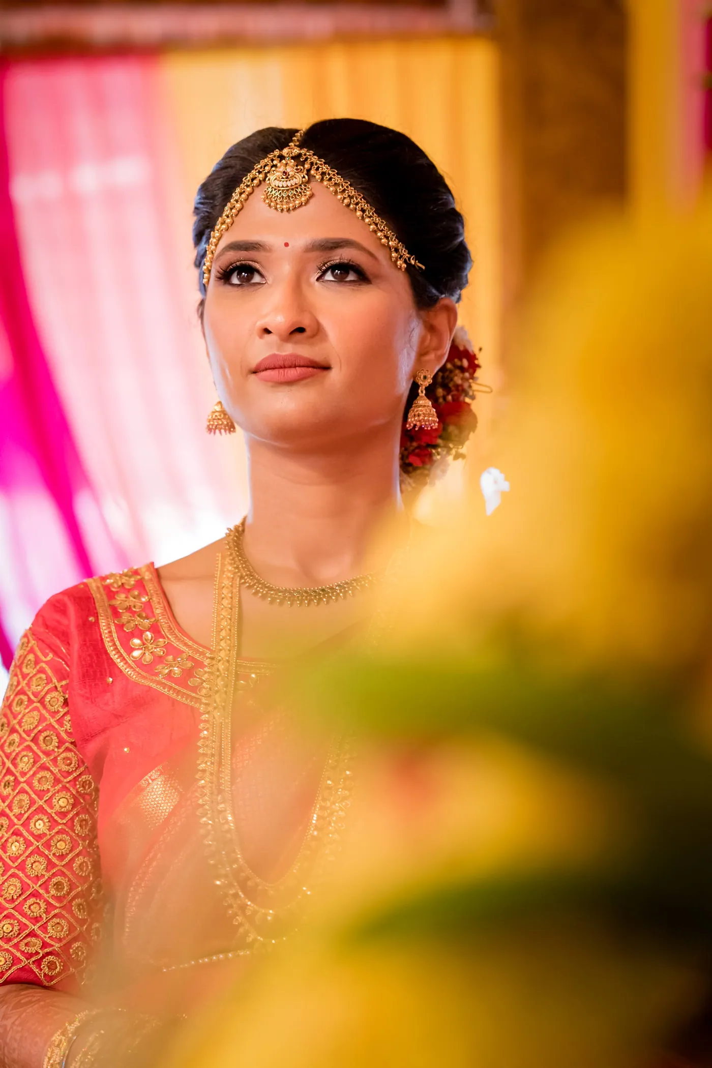 Medha in an orange blouse looks upward with a serious gaze between pink drapes and blurred yellow flowers