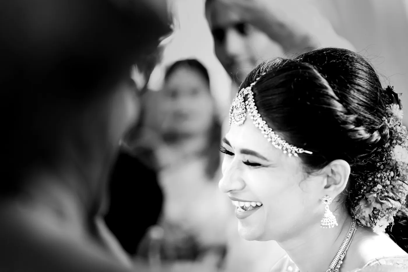 Black and white of Medha laughing warmly in a gold headpiece, a blurred figure in front of her